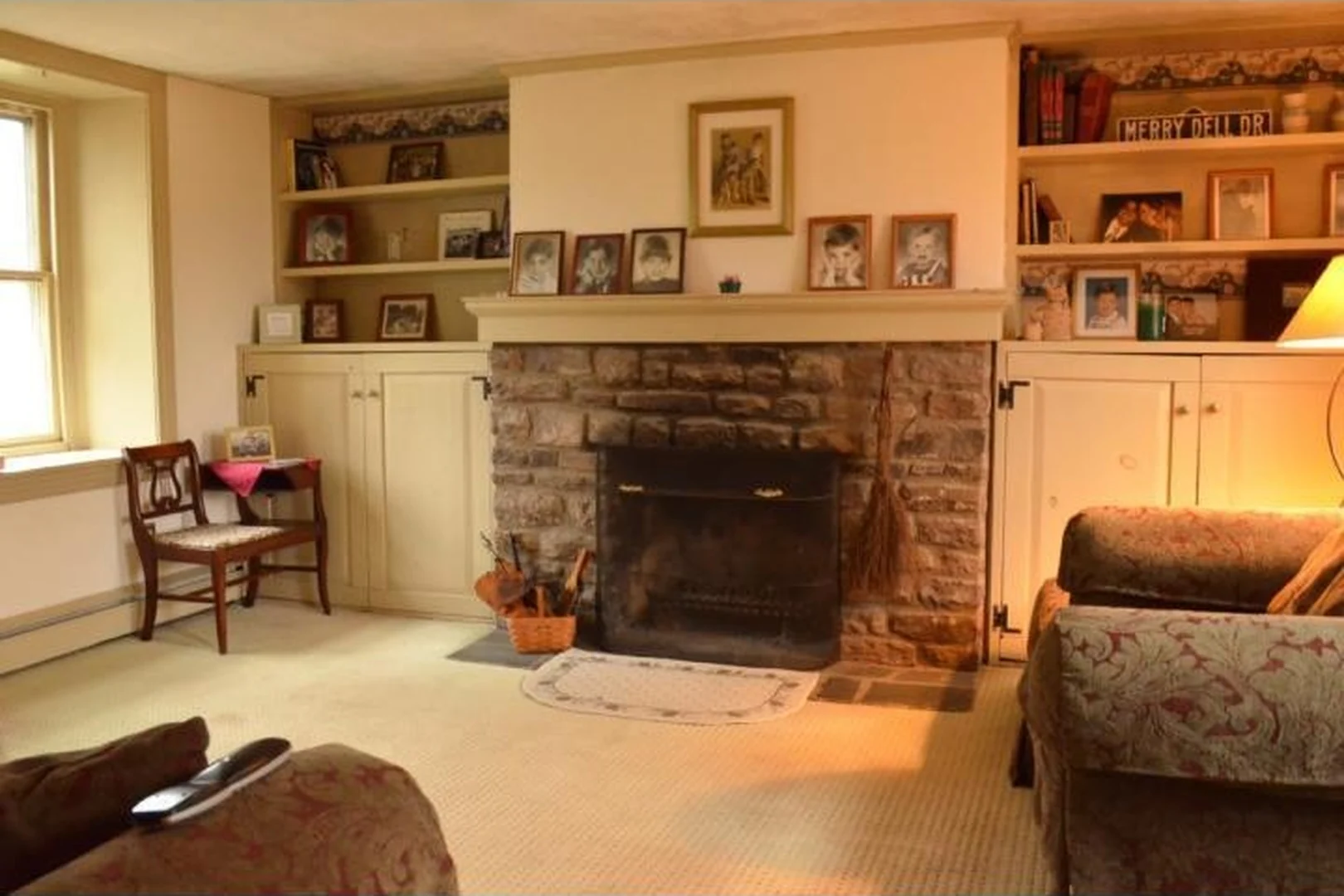 Bucks County living room before renovation, with wall-to-wall carpet, a soot-stained stone fireplace, builder-grade painted built-ins crowded with framed photos, and dated upholstered furniture
