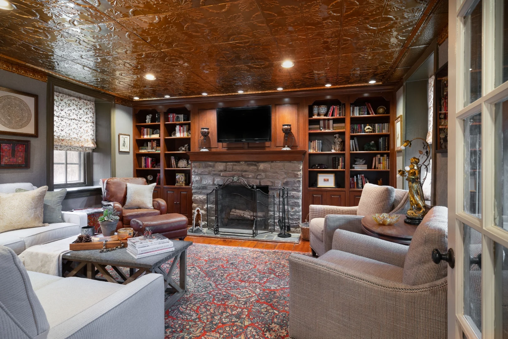 Bucks County living room after renovation, viewed from the doorway, with a white sectional, houndstooth swivel chairs, a leather club chair, a zinc-top coffee table, and the fireplace beneath the tin ceiling