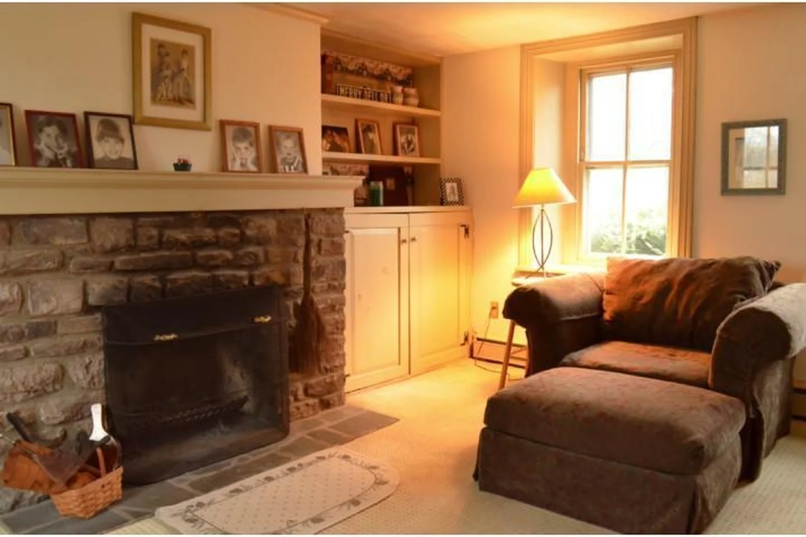 Bucks County living room before renovation, viewed at an angle, with beige walls, carpet, a bulky brown microfiber chair and ottoman, and a brass floor lamp beside the stone fireplace