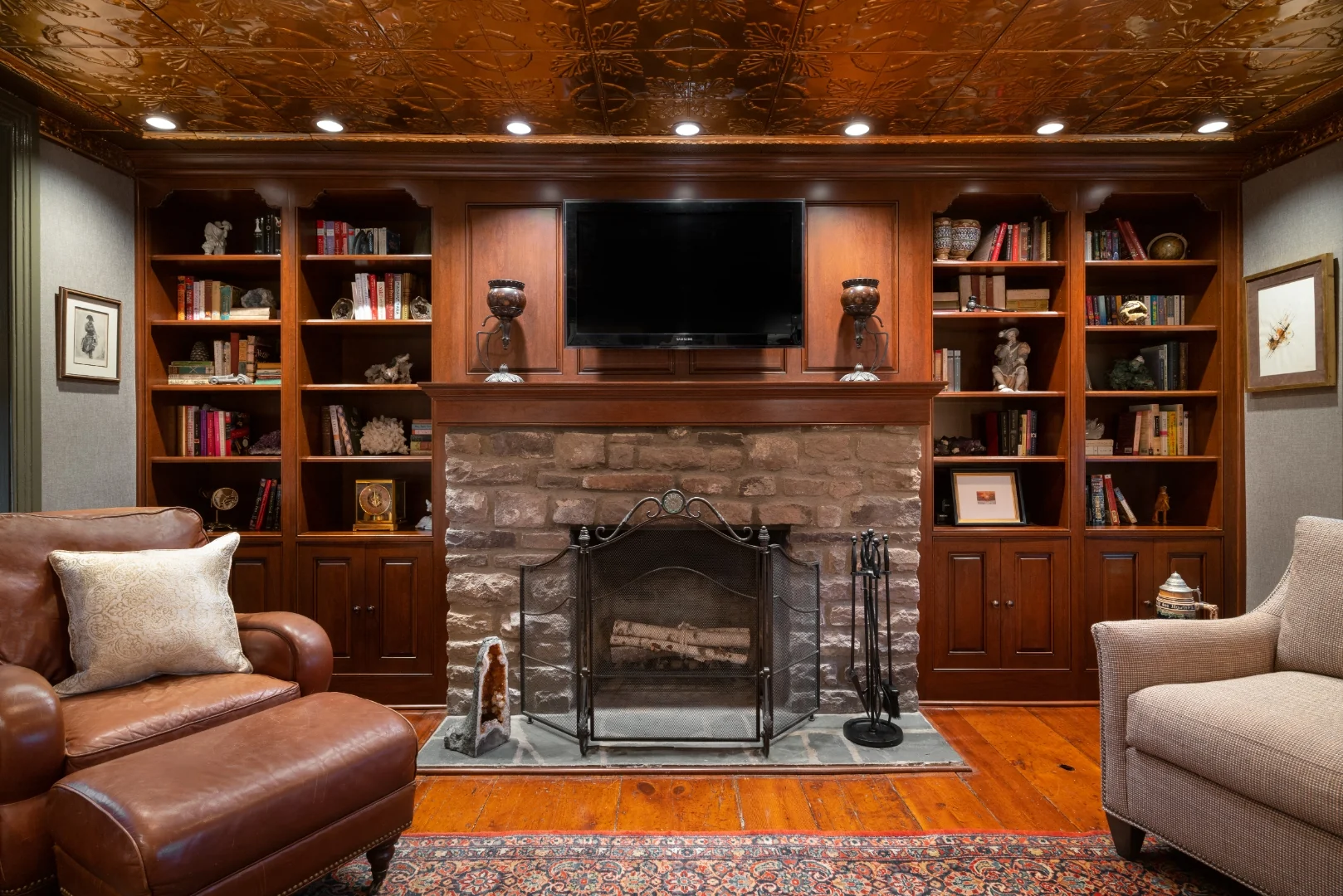 Restored Bucks County historic living room with an antique pressed-copper tin ceiling, custom cherry built-in bookcases flanking a stone fireplace, a leather club chair, and an oriental rug on wide-plank wood floors