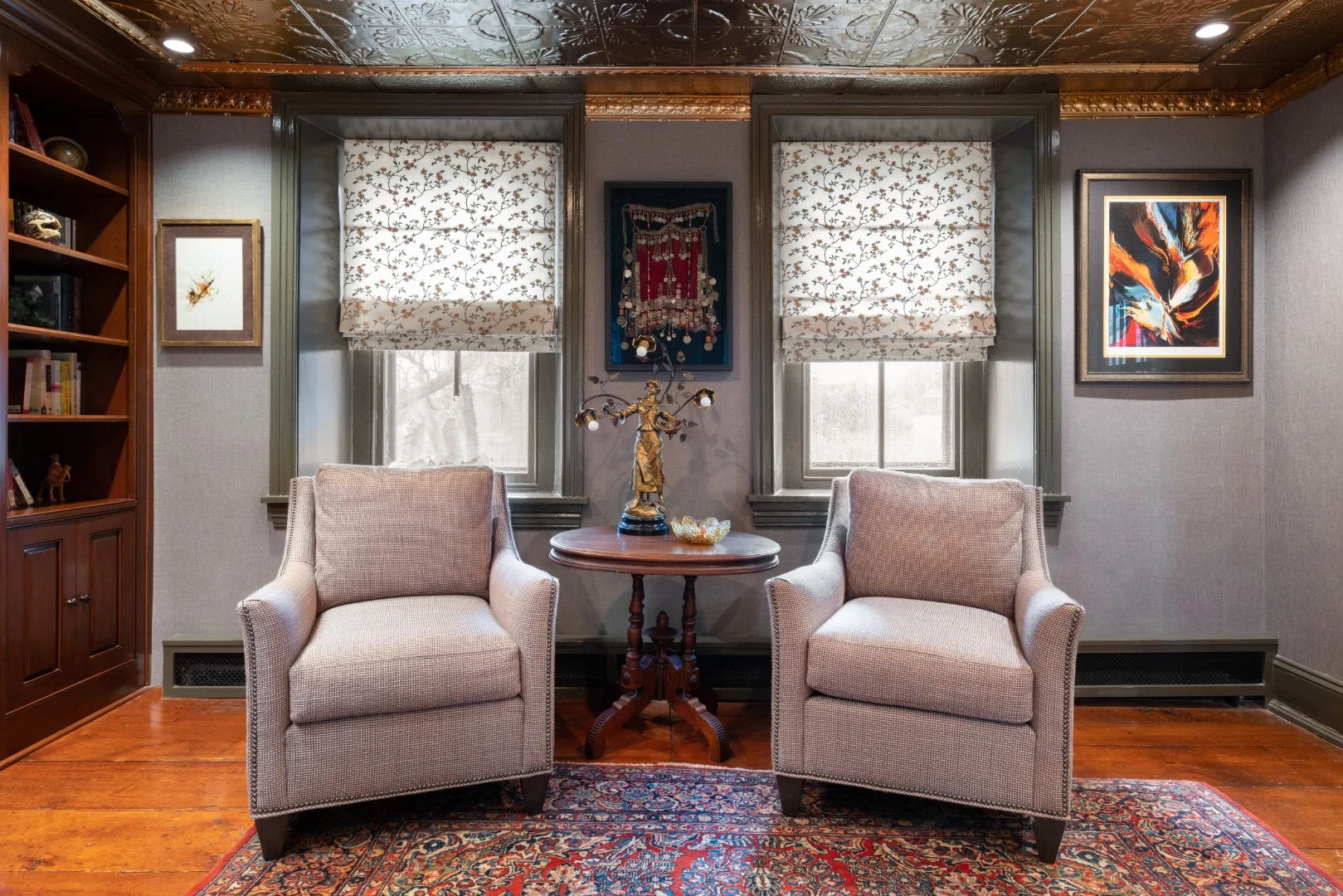 A pair of houndstooth armchairs flanking a carved Victorian parlor table and gilt figural lamp between two windows with floral roman shades in the Bucks County living room