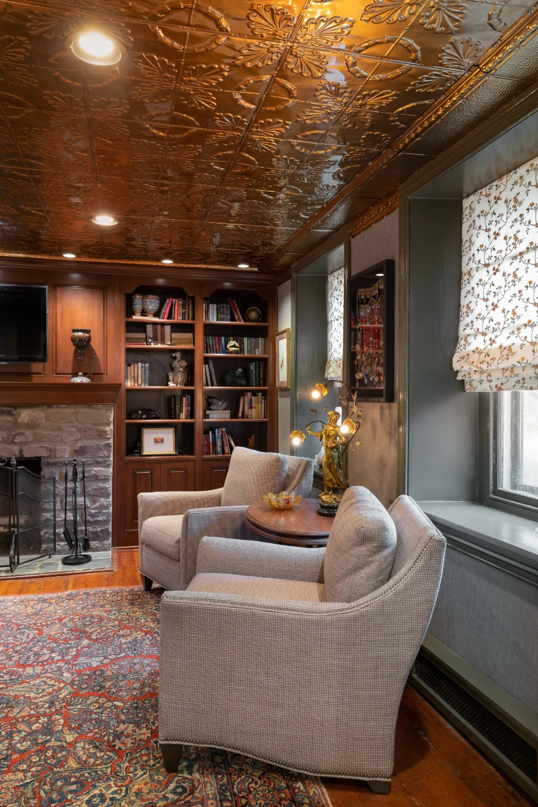 Reading corner with cherry bookcases, the stone fireplace and mounted TV, houndstooth chairs around a round table, and a window seat beneath the pressed-tin ceiling in the Bucks County living room