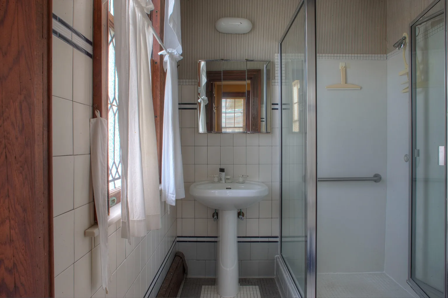 Cape May bathroom before renovation with a white pedestal sink, dated floral wallpaper, red-trimmed wall tile, and a basic framed mirror