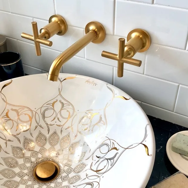 Detail of the artisan white vessel sink with a gold filigree pattern and a brushed-gold wall-mount cross-handle faucet on a black lacquer vanity in the Cape May chinoiserie bathroom