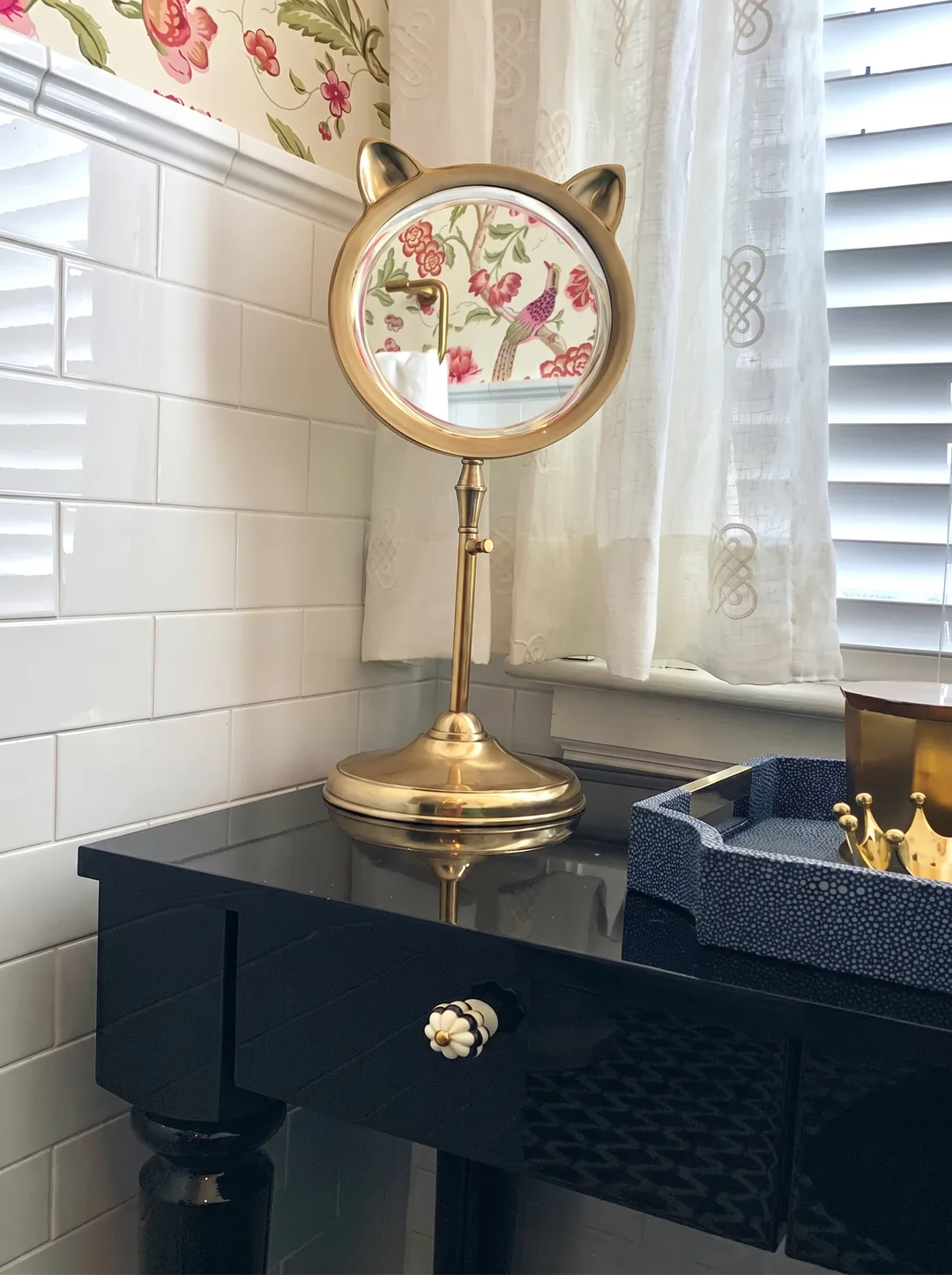 Brass cat-ear adjustable vanity mirror on the black lacquer table with a ceramic floral knob, white subway tile, and a curtained window in the Cape May chinoiserie bathroom
