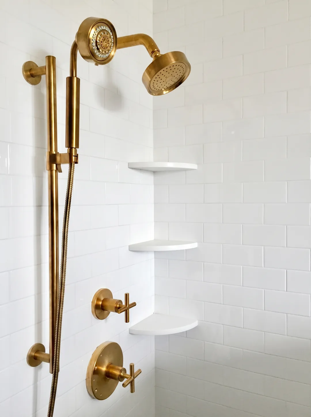 Detail of the polished brass rainfall shower head, handheld wand, and cross-handle valves with white corner shelves on white subway tile in the Cape May chinoiserie bathroom