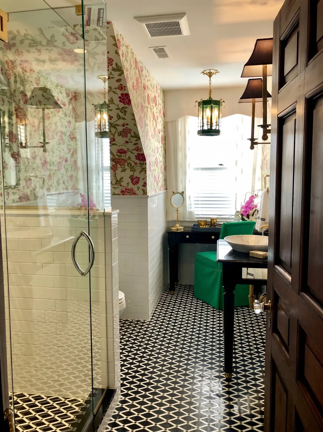 View into the Cape May chinoiserie bathroom from the doorway showing the frameless glass shower, floral wallpaper, black lacquer vanity, green velvet chair, and geometric floor tile