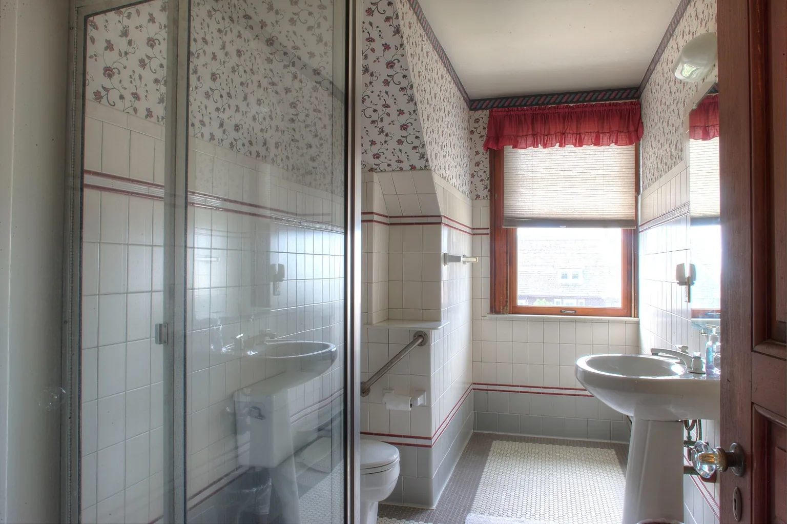 Cape May hall bathroom before renovation with dated mauve floral wallpaper, red-striped trim, a white pedestal sink, a builder-grade corner glass shower stall, and hex floor tile
