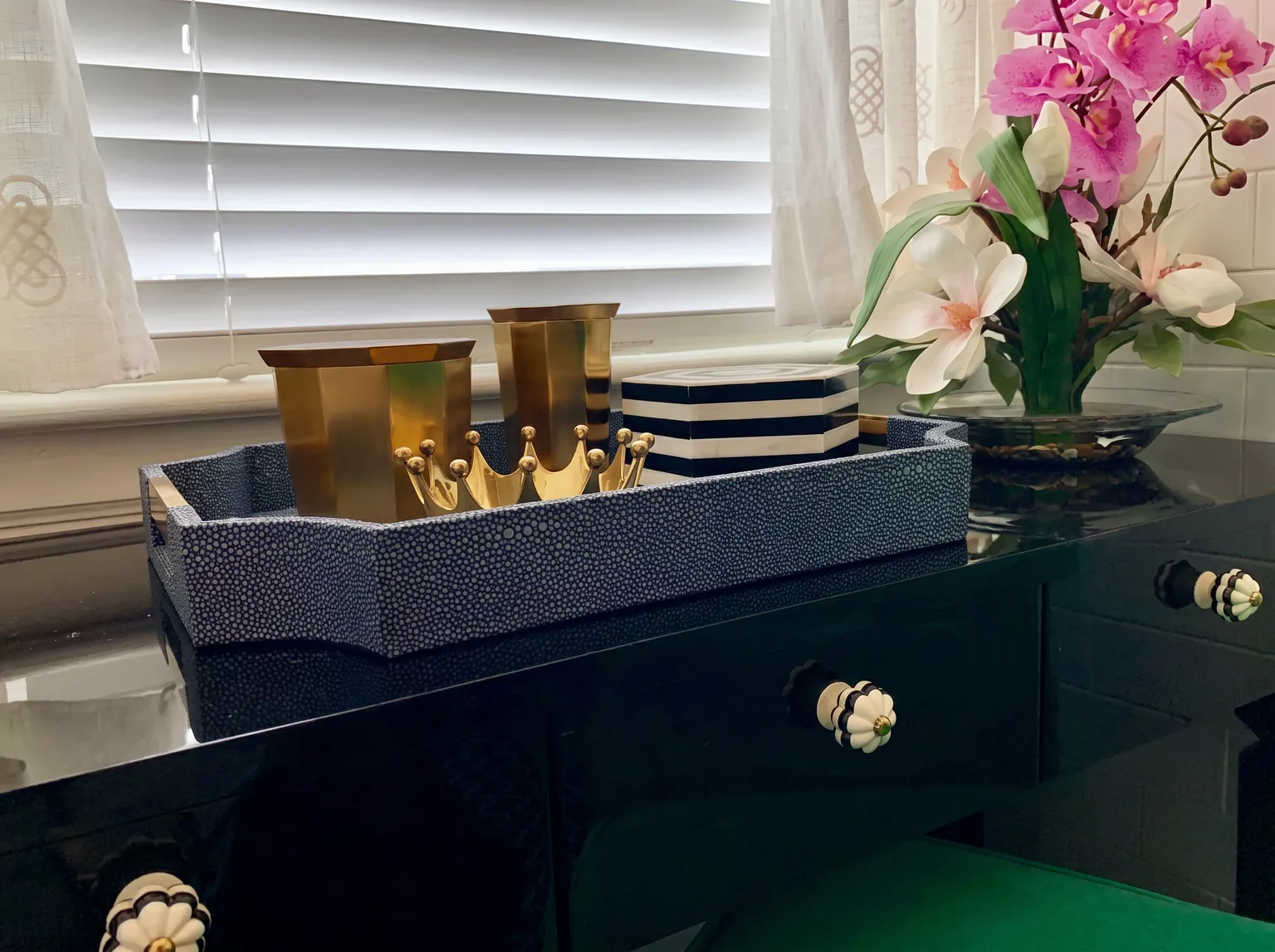 Styled vignette on the black lacquer vanity with a navy shagreen tray holding brass cups and a gold crown ornament, a striped box, and a pink orchid in the Cape May chinoiserie bathroom