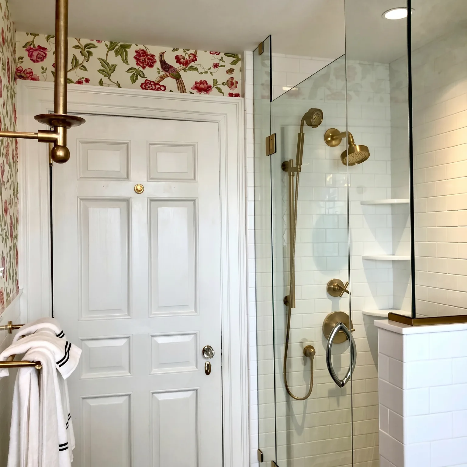Cape May chinoiserie hall bathroom after renovation with a frameless glass corner shower, brass fixtures, white subway tile, a crisp white paneled door, and floral wallpaper above