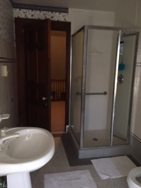 Cape May hall bathroom before renovation with a dated framed corner shower stall, a dark wood paneled door, and floral-papered walls