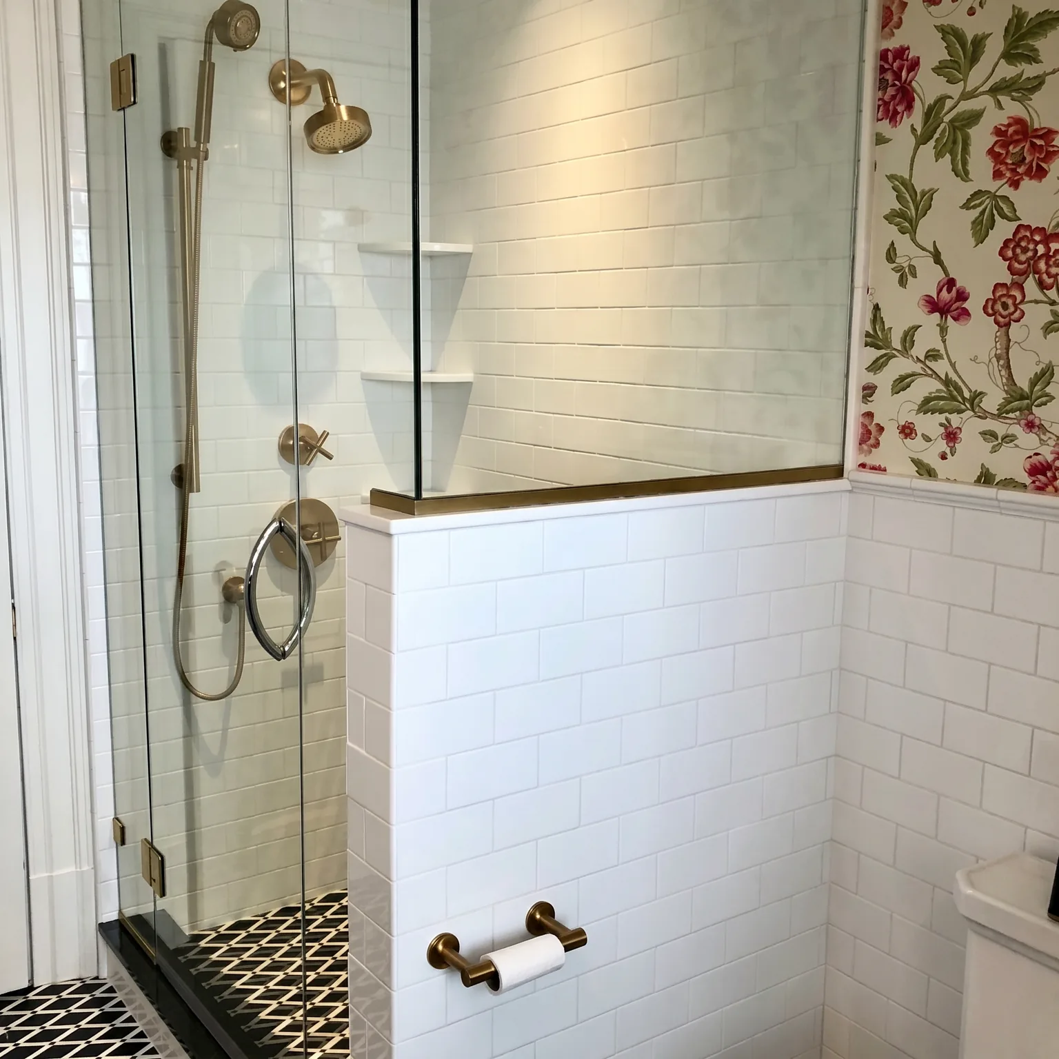 Frameless glass shower corner with brass and chrome fixtures, white subway wainscot, floral wallpaper, and a brass toilet-paper holder in the Cape May chinoiserie bathroom