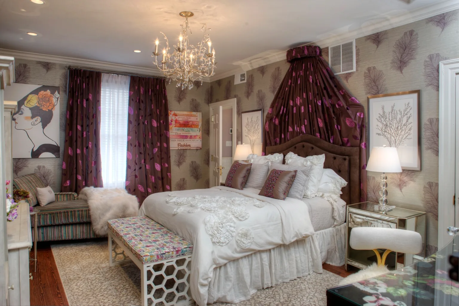 Glamorous Cape May ensuite bedroom with a velvet upholstered bed, draped fabric crown, crystal chandelier, and feather-motif grasscloth wallpaper