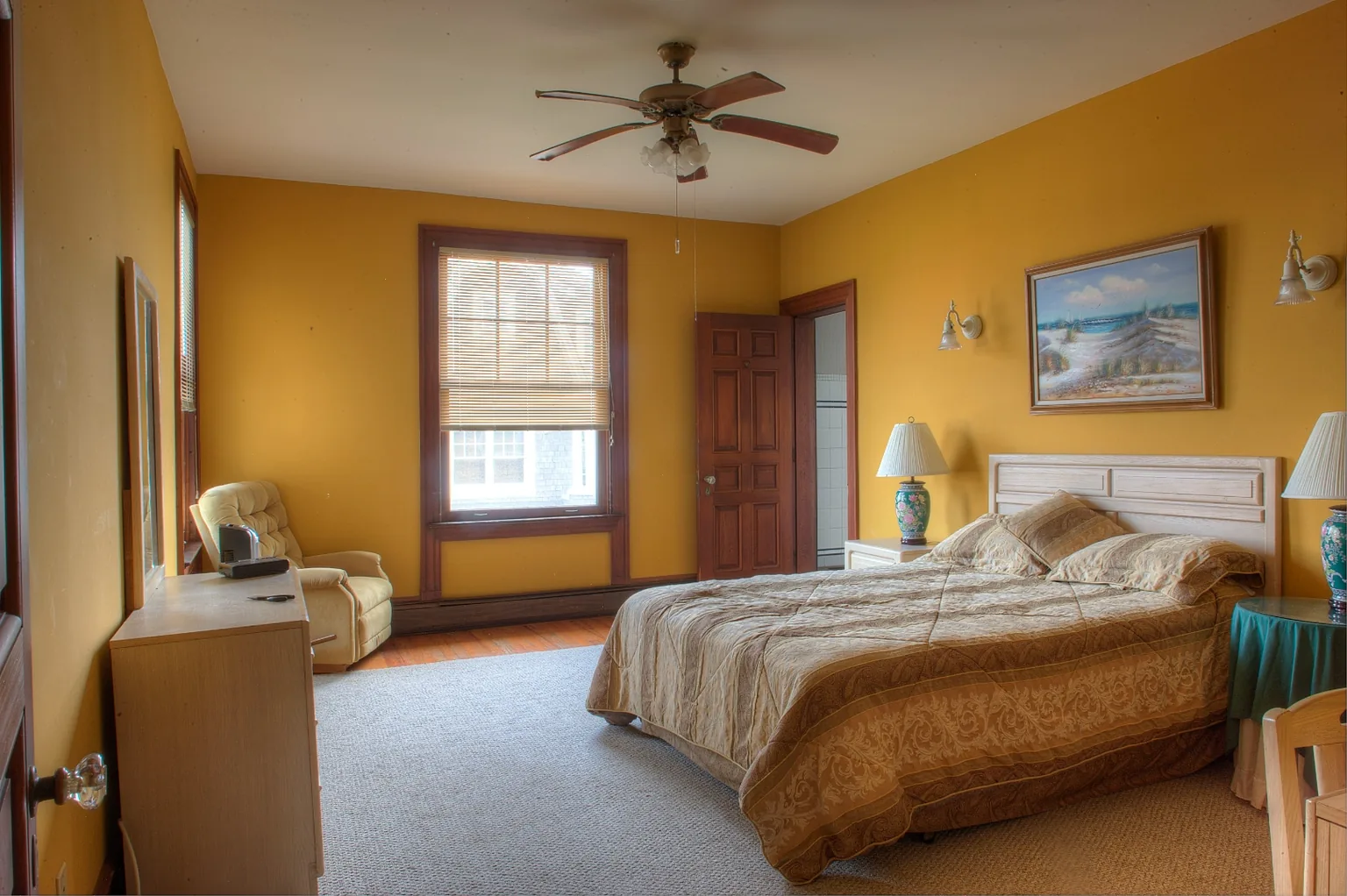 Cape May bedroom before renovation with golden-yellow walls, a dated ceiling fan, light oak headboard, and a beach painting