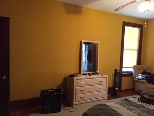 Cape May bedroom dresser wall before renovation with bare yellow walls, a laundry basket, and a small white dresser