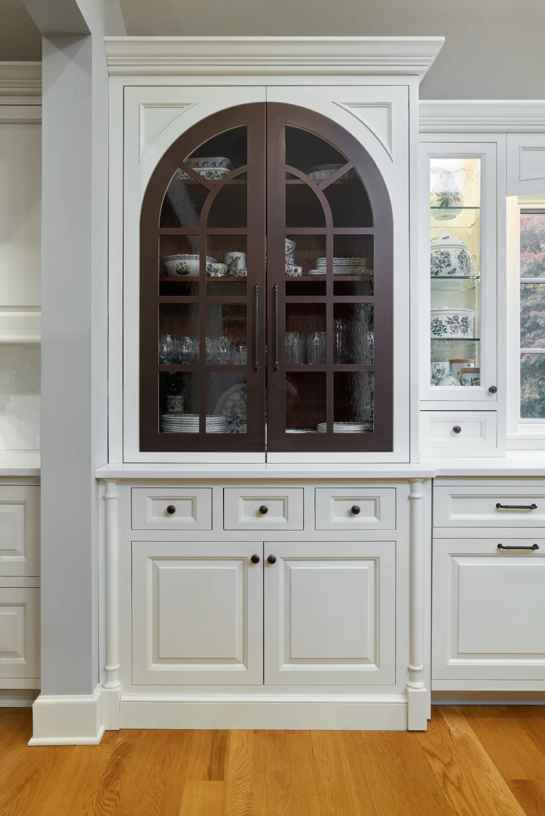 Arched glass display hutch with a burgundy-stained interior holding botanical china in a Chester, PA kitchen