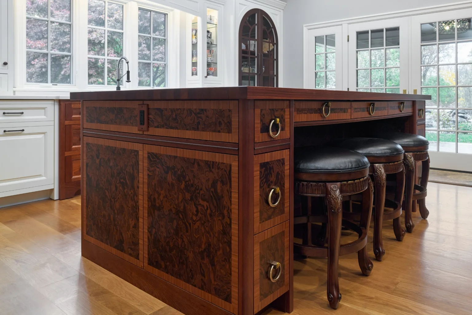 Burlwood kitchen island with a seating overhang and black leather carved-wood swivel stools beneath an arched glass display cabinet