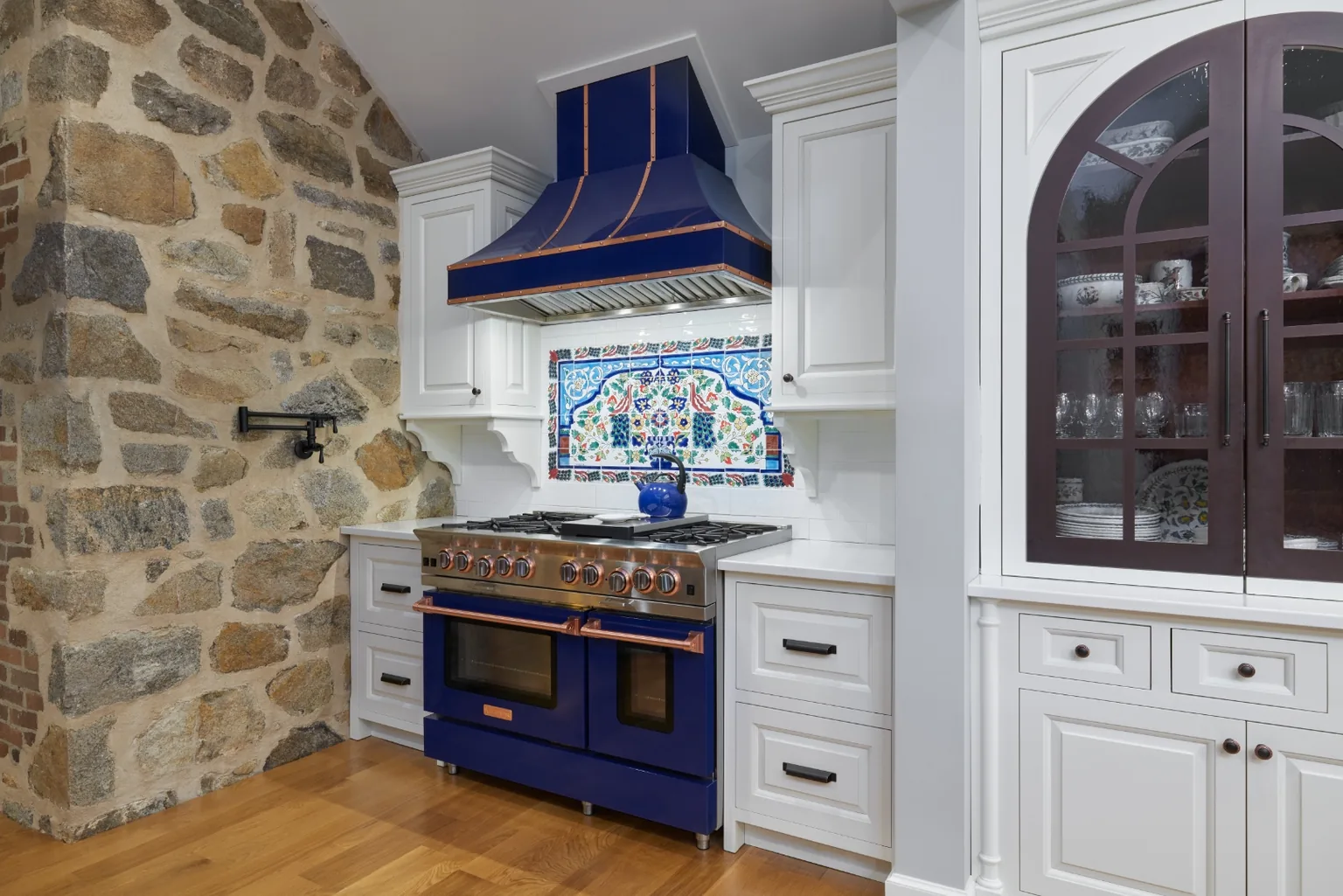 Cobalt blue and copper range set against an exposed natural stone wall with a hand-painted tile mural backsplash in a Chester, PA kitchen