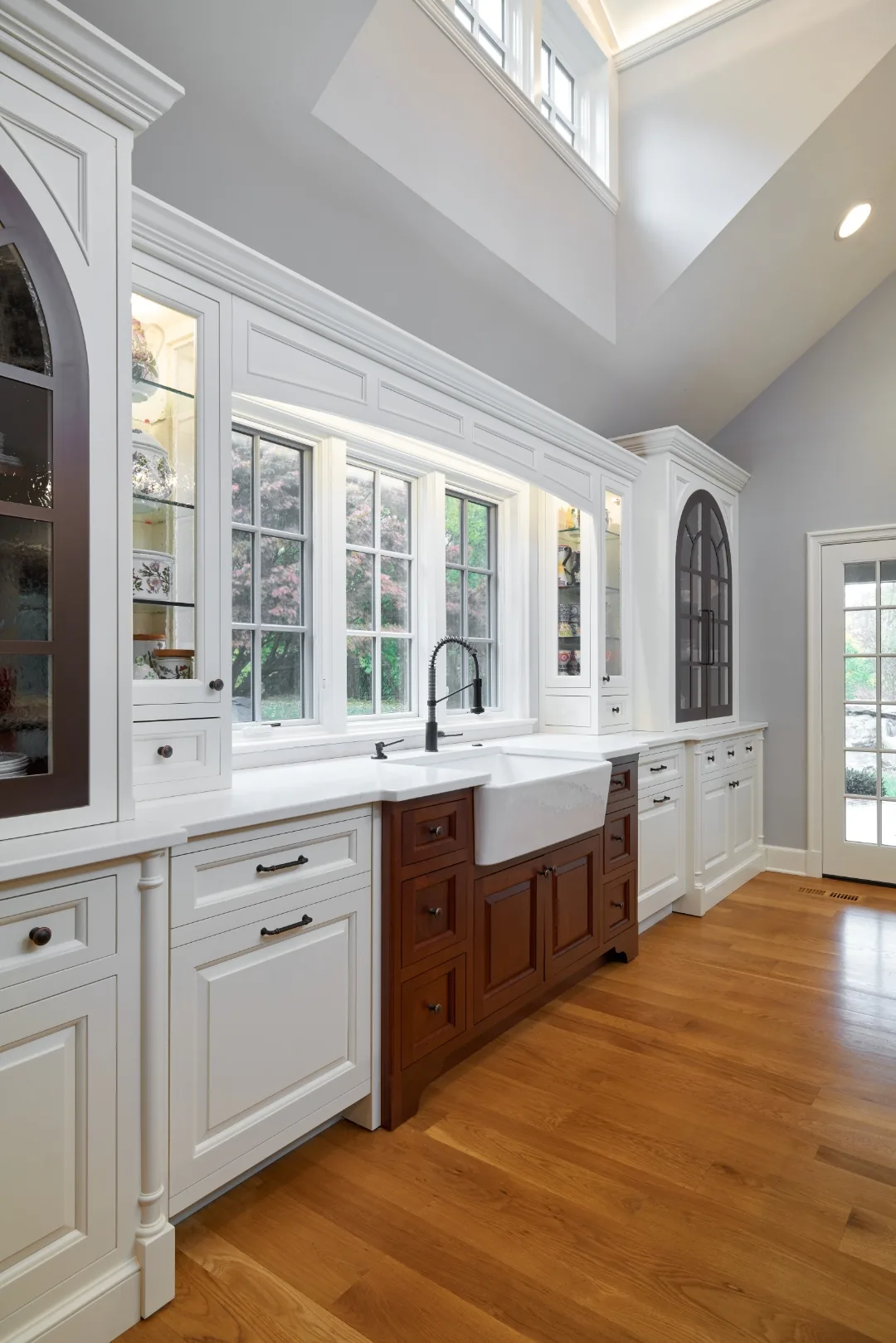 Window wall with a farmhouse apron sink, white and cherry cabinetry, and arched glass-front cabinets in a Chester kitchen