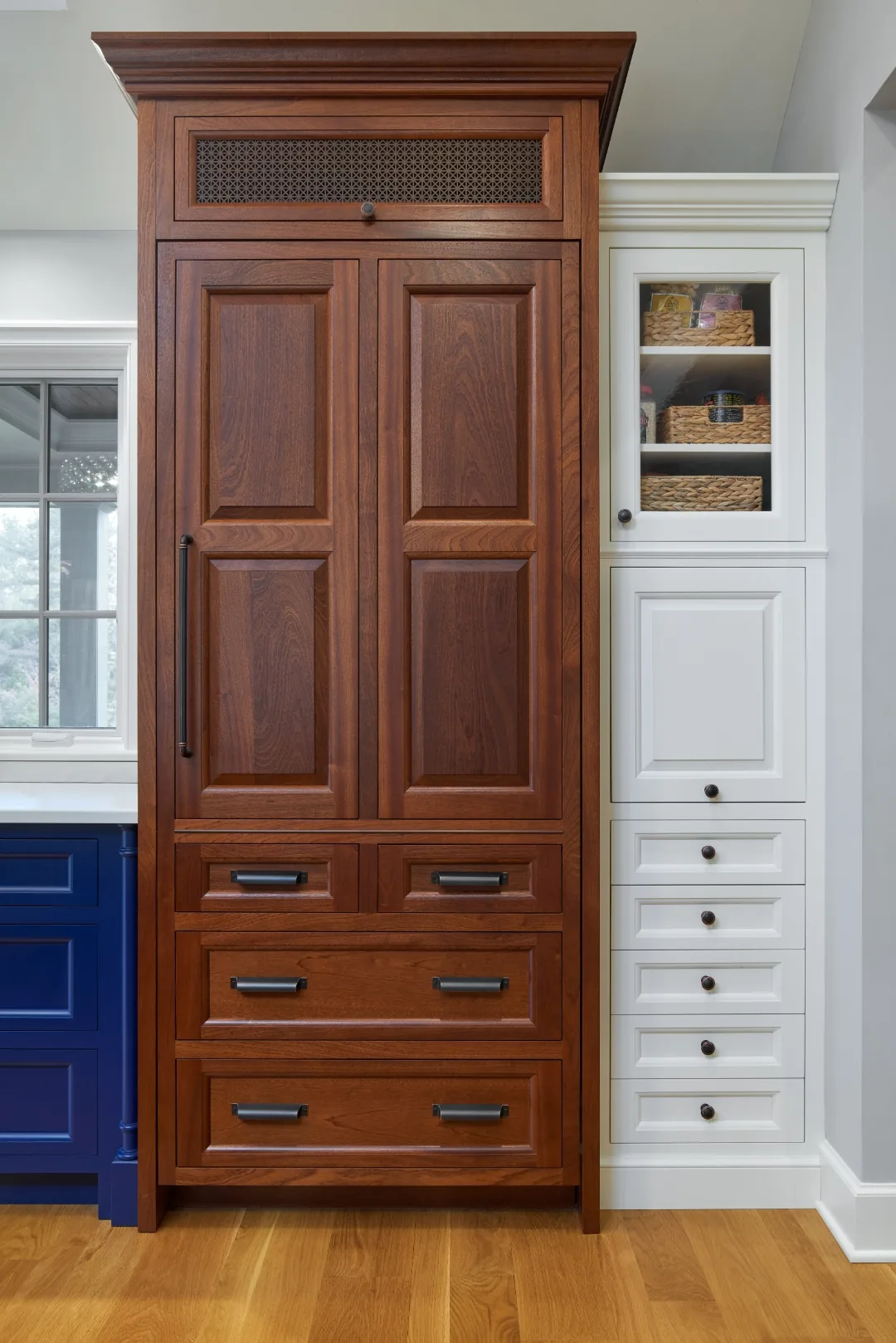 Paneled cherry refrigerator surround with a pierced-metal grille flanked by navy cabinetry and a white pantry tower