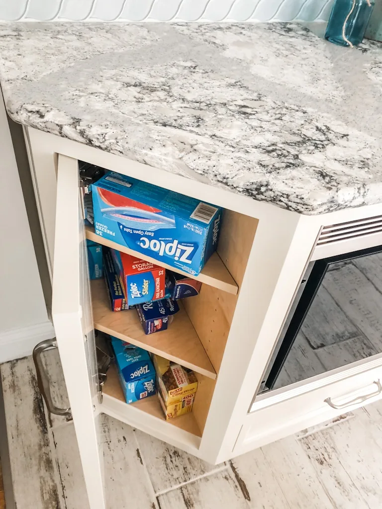 Angled corner base cabinet with adjustable shelves storing boxed wraps and bags beneath a speckled granite counter and blue tile in the Churchville kitchen