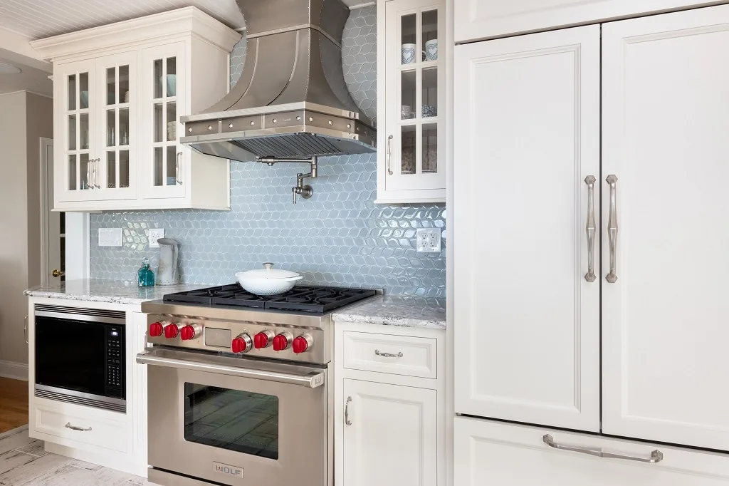 Angled view of the Churchville kitchen appliance wall showing the Wolf range, built-in microwave, panel-ready refrigerator, glass uppers, and blue hexagon tile backsplash