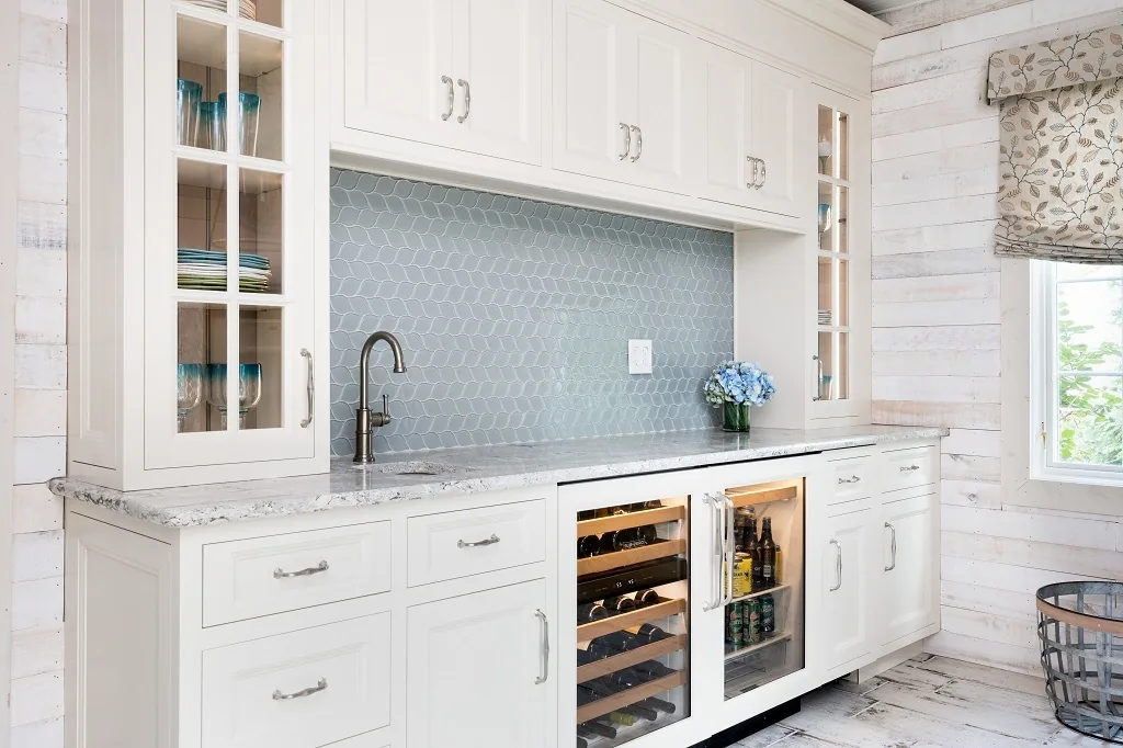 Angled view of the Churchville beverage center with glass-front cabinets, blue hexagon tile, a marble counter, a built-in wine and beverage fridge, and a window with floral shade