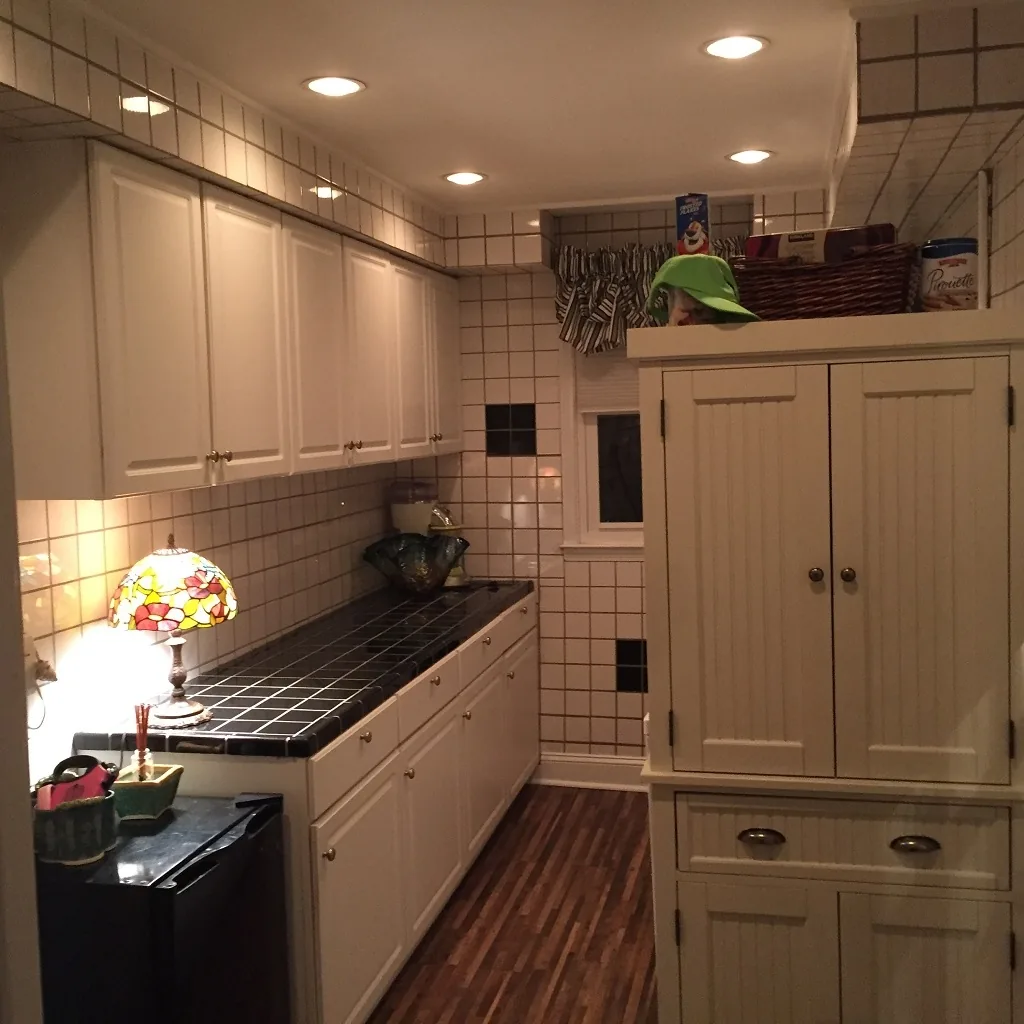 Churchville beverage area before renovation with dated white grid tile walls, a black grid-tile countertop, a beadboard cabinet, a stained-glass lamp, and a small window