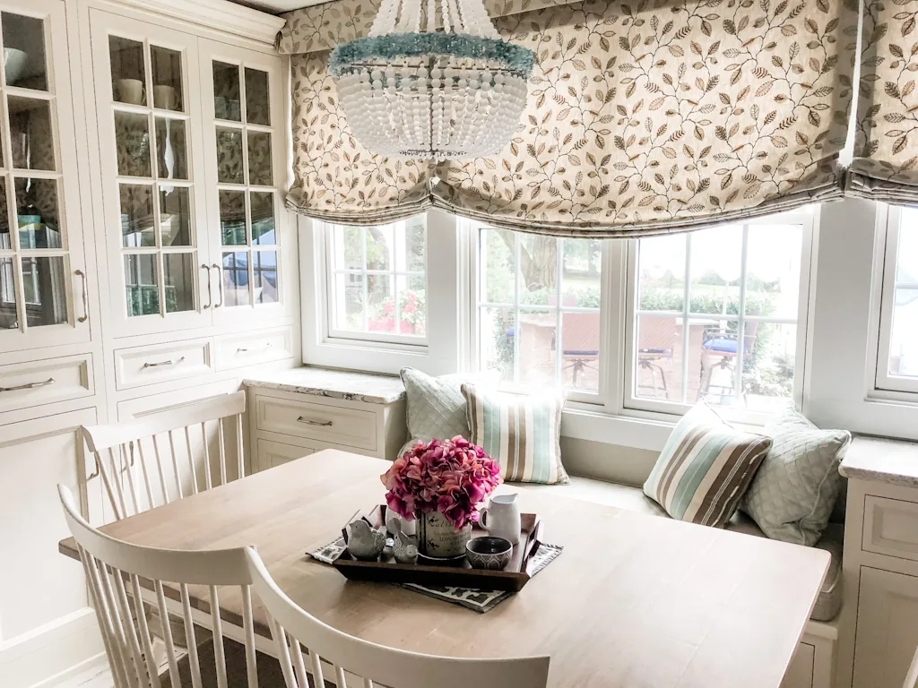 Churchville breakfast nook with a light wood table, white spindle chairs, a built-in glass hutch, a bayed window seat with aqua and striped pillows, and a beaded chandelier