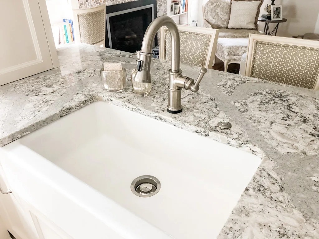 White farmhouse apron sink with a brushed-nickel pull-down faucet set in a speckled granite island counter, with the breakfast area beyond in the Churchville kitchen