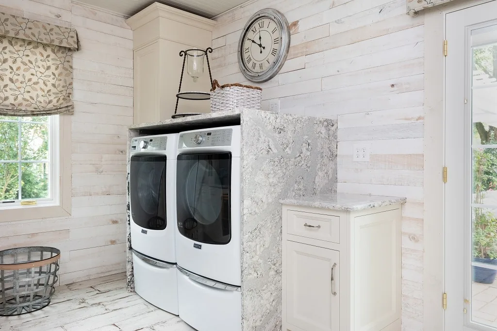 Hidden laundry area in the Churchville kitchen with a white front-load washer and dryer concealed behind a marble surround, whitewashed shiplap walls, and a wall clock