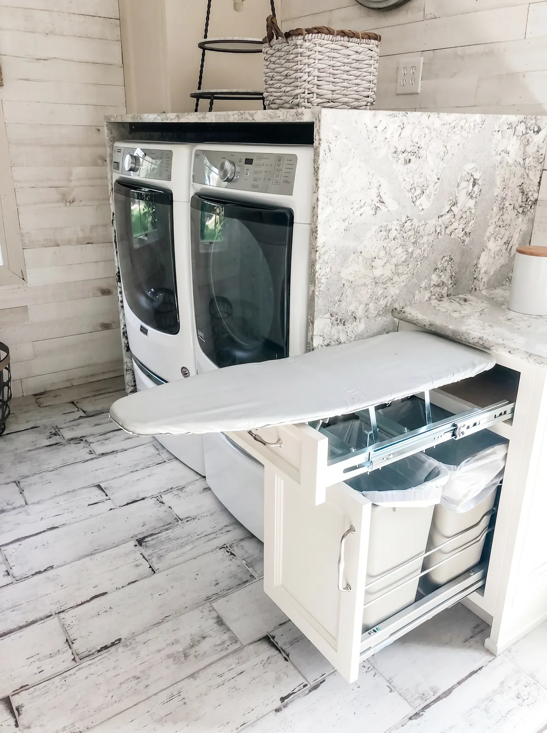 Custom pull-out ironing board and hidden pull-out trash cabinet beside a stacked white washer and dryer with whitewashed shiplap walls and a marble counter in the Churchville kitchen