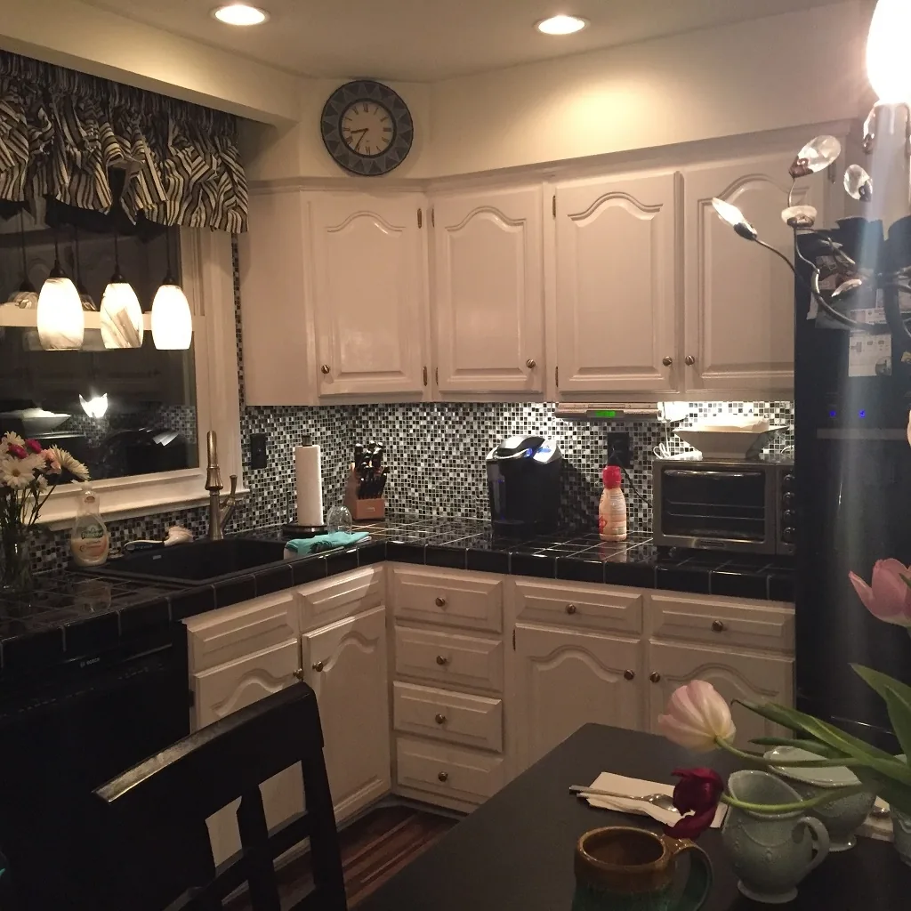 Churchville kitchen before renovation with dated white raised-panel cabinets, black tile countertops, a dark mosaic backsplash, a zebra-print valance, black appliances, and a wall clock