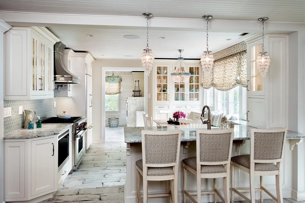 Churchville beach house chic kitchen overview with white custom cabinetry, a center island with patterned counter stools, three beaded chandeliers, a beadboard ceiling, glass-front upper cabinets, and whitewashed plank floors