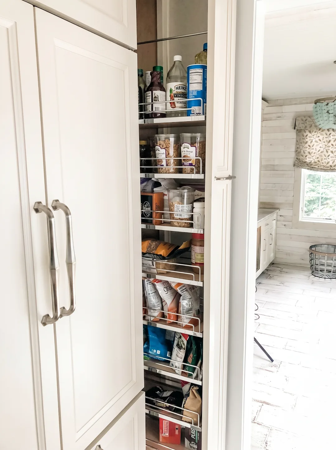 Tall narrow pull-out wire pantry organizer stocked with dry goods beside a panel-ready refrigerator, with the whitewashed kitchen visible through the doorway in the Churchville kitchen