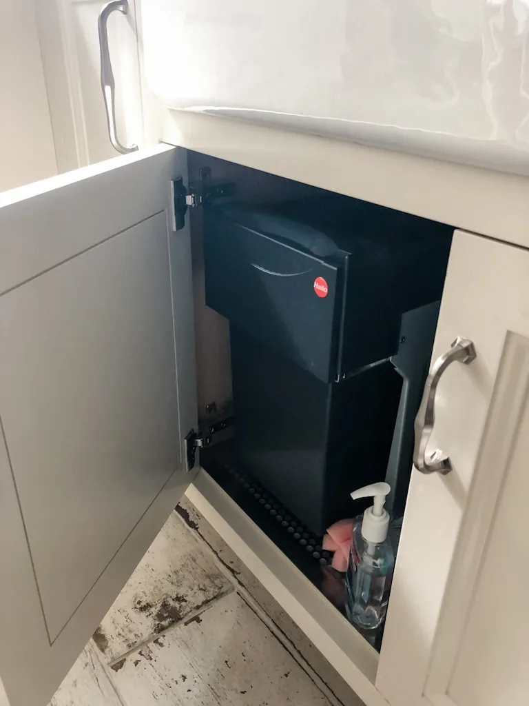 Concealed pull-out waste bin in the cabinet beneath the white apron-front sink with whitewashed plank flooring in the Churchville kitchen
