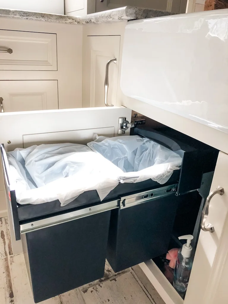 Dual pull-out trash and recycling bins concealed beneath the white farmhouse apron sink in the Churchville kitchen