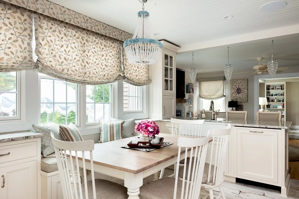 Churchville kitchen breakfast area with a light wood table and white spindle chairs, a beaded chandelier, floral roman shades, a window seat, and the island prep sink beyond