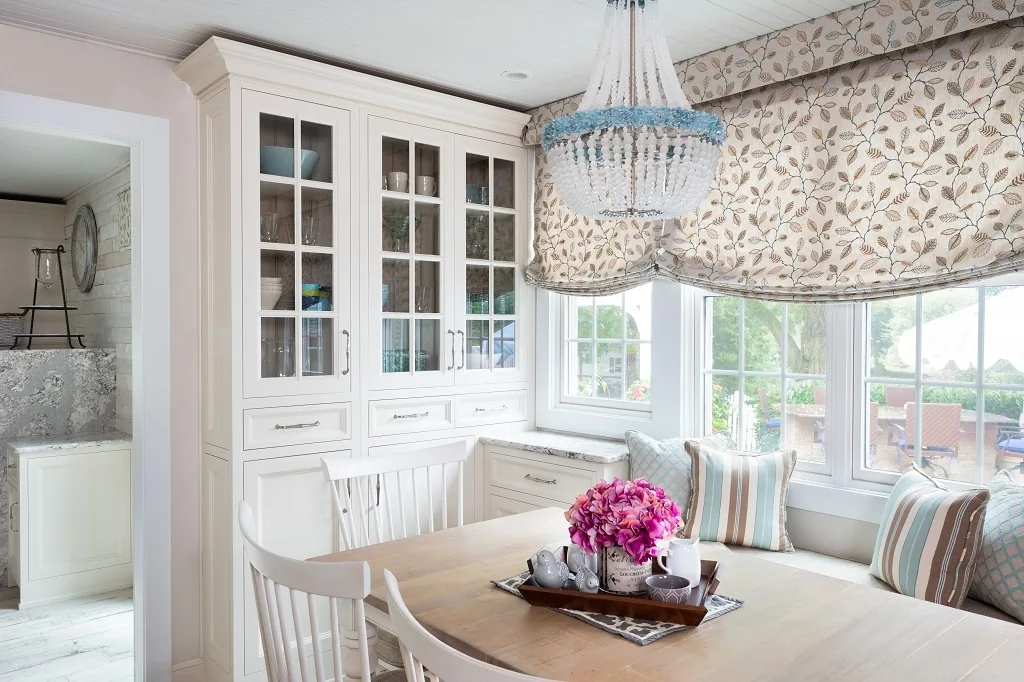 Built-in glass-front display hutch beside the window-seat breakfast nook with a beaded chandelier, floral roman shades, striped pillows, and a light wood table in the Churchville kitchen