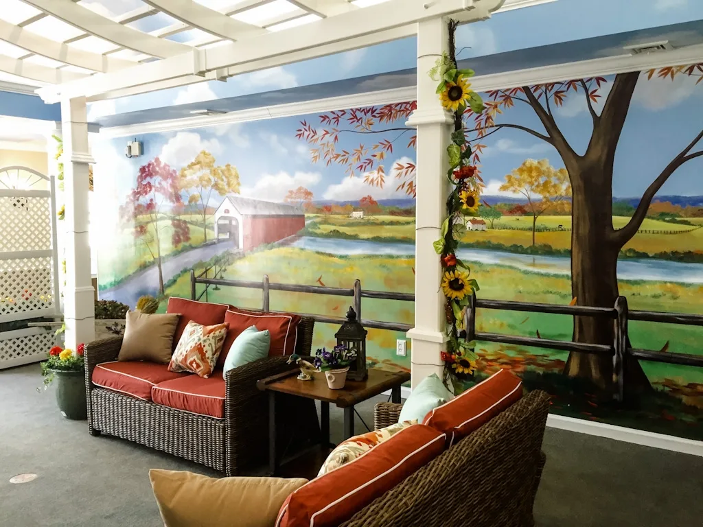Immersive memory care sensory room with painted sky ceiling, faux stone wall, pergola, and hand-painted countryside mural