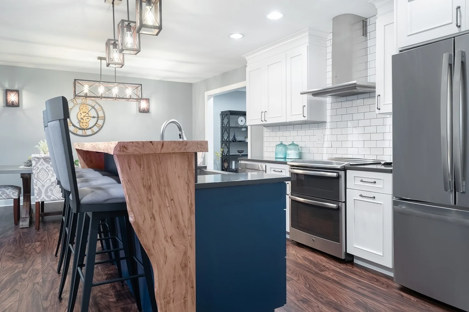 Holland kitchen cooking wall after renovation, with white shaker cabinets, a white subway tile backsplash, dark quartz countertops, a stainless double-oven range under a stainless hood, a stainless French-door refrigerator, and a navy island with a live-edge waterfall wood bar