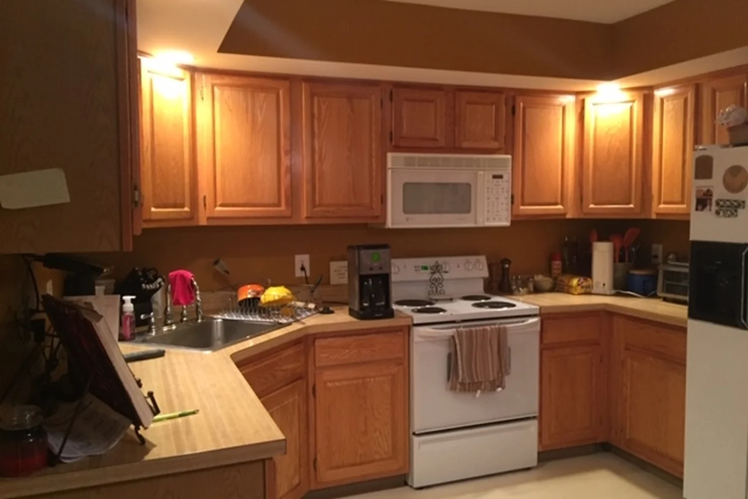 Holland kitchen cooking wall before renovation, with dated honey-oak cabinets, beige laminate countertops, a white electric range, an over-the-range microwave, white appliances, and gold-brown walls