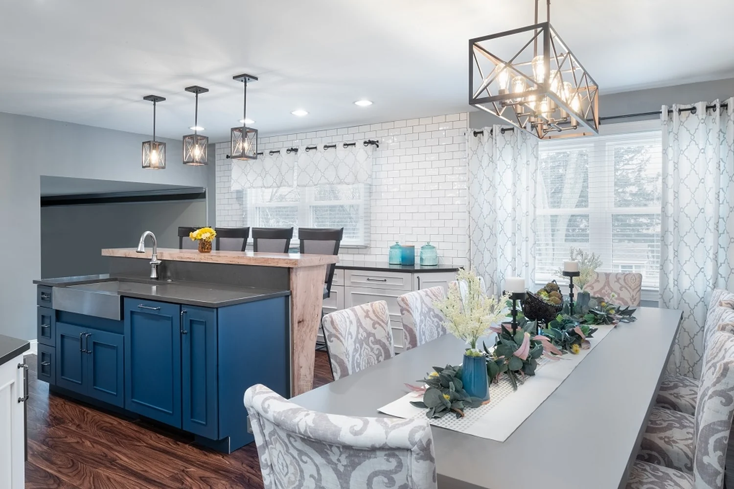 Holland eat-in dining area after renovation, with a long gray table and patterned upholstered chairs, a navy island with a stainless farmhouse apron sink and live-edge waterfall bar, a white subway tile wall, layered window treatments, and a geometric pendant chandelier