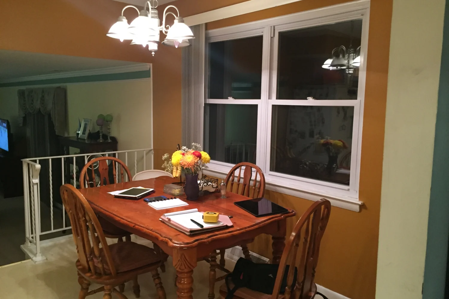 Holland eat-in area before renovation, with gold-brown walls, an oak pedestal table and spindle chairs, a dated white chandelier, and a double-hung window with no treatments