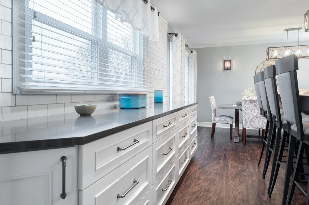 White shaker base cabinetry and buffet run with dark quartz countertop and oil-rubbed bronze pulls beneath subway tile and bright windows, looking toward the eat-in dining table in the Holland mix and match kitchen