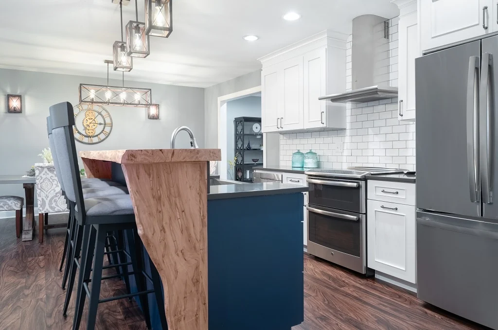 Side view of the navy island and live-edge waterfall wood bar with gray barstools, the white subway tile cooking wall, a stainless double-oven range and hood, a stainless French-door refrigerator, and walnut floors in the Holland kitchen