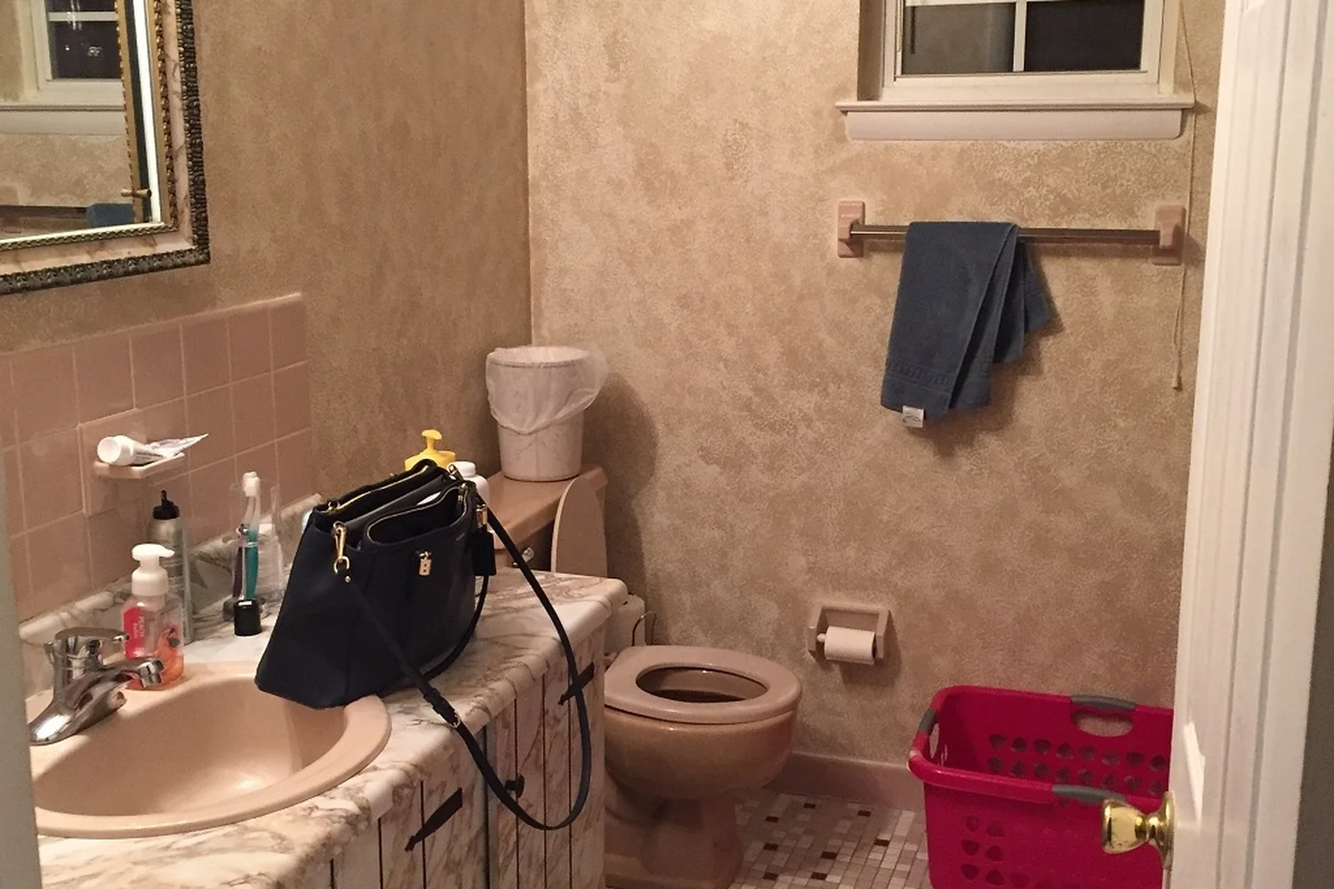 Holland bathroom before renovation, viewed from the doorway, with a dated faux-marble vanity and pink sink on the left, beige faux-finish walls and a wallpaper border, a beige toilet, and a small window with old blinds