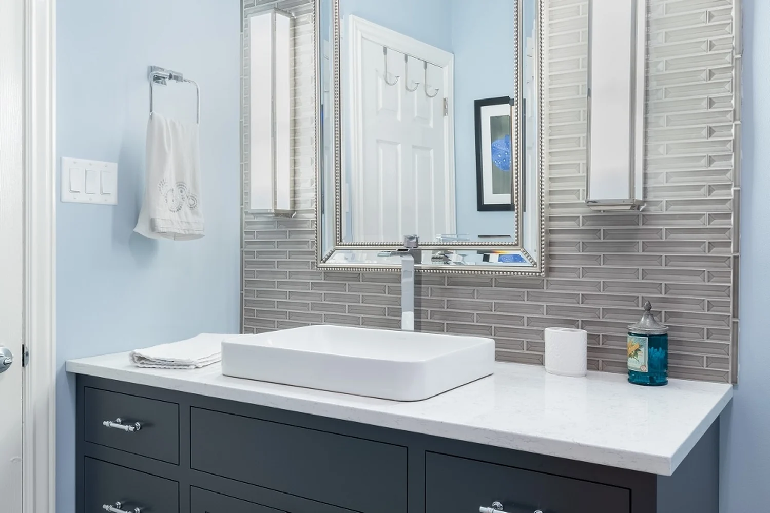 Holland bathroom vanity after renovation, a custom navy Nina Green Signature Series cabinet with a white quartz countertop, a rectangular vessel sink, a polished chrome waterfall faucet, a beaded silver mirror, and a shimmering linear glass tile accent wall