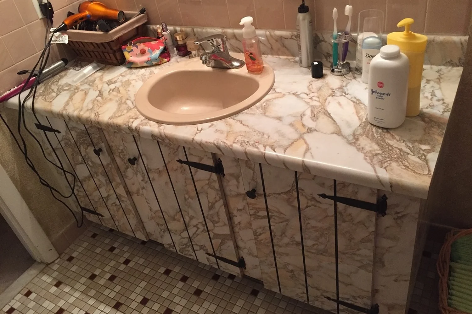 Holland bathroom vanity before renovation, with a cluttered faux-marble countertop, a pink oval drop-in sink, dated faux-marble cabinet doors with black hinges, and mosaic floor tile