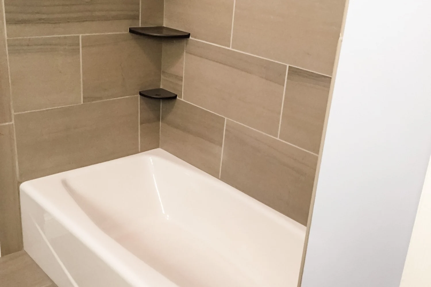 Holland bathroom tub after renovation, with a clean white tub, large-format wood-look porcelain wall tile, three floating dark corner shelves, and modern chrome fixtures